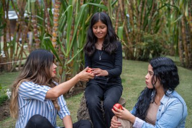 Annesi kızına taze kırmızı bir elma sunarken kız kardeşi yakınlarda oturuyor, aile anlarının tadını çıkarıyor ve yemyeşil bir bahçede sağlıklı bir şekilde yemek yiyor.