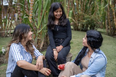 Aralarında neşeli bir kızı ve aile fertlerinin de bulunduğu üç nesil kadın, yemyeşil bir bahçede çimlerin üzerinde oturmuş kırmızı elmaları tutarken kahkahalarını ve mutluluklarını paylaşıyorlar.
