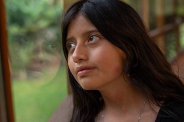 Camdan dışarı bakan uzun siyah saçlı genç bir kız. Yüzü kısmen cama yansıyor. Düşünce ve düşüncelere dalmış.