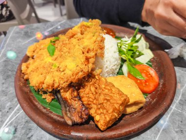 Delicious Indonesian smoked fish, rice, tempeh, tofu, and fresh vegetables on a traditional plate.