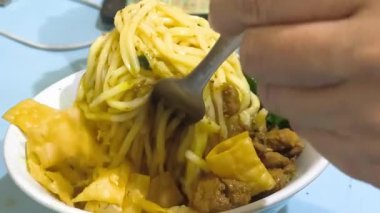 Mie Ayam or Indonesian Chicken Noodle close up. Noodles with seasoned chicken, mustard greens, and fried wonton crackers, being eaten with a fork and spoon. Asian street food.
