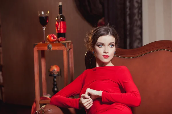 Porträt eines schönen Mädchens mit dunklen Haaren und blauen Augen in einem roten Kleid auf einem braunen Sofa mit einem Glas Rotwein und Granatapfel — Stockfoto