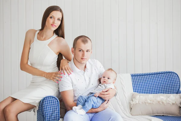 Una familia joven con un pequeño bebé agradable y dulce sentado en el sofá en un interior brillante —  Fotos de Stock