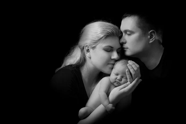 Lycklig familj, unga föräldrar som innehar en nyfödd bebis i famnen och försiktigt kramade honom, svartvitt foto på en svart bakgrund . — Stockfoto