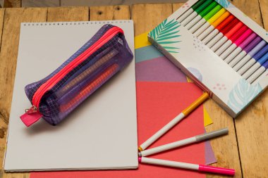 Okul ve sanat malzemeleri ahşap yüzeye dizilmiş, mor ağ kalem kutusu, çizim defteri, renkli kağıt ve yaratıcı çalışma alanındaki gradyan işaretleyici kutu içeriyor..