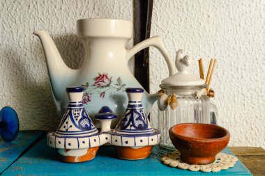 Çiçek desenli beyaz seramik bir çaydanlık, iki küçük mavi ve beyaz tagine tenceresi ve kuş heykelciği ve sazlık difüzyon çubuklarıyla süslenmiş beyaz kapaklı klasik bir cam kavanoz içeren bir yaşam düzenlemesi..