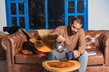 Gitar ve kedinin yanında metal çaydanlıktan çay dolduran kişi