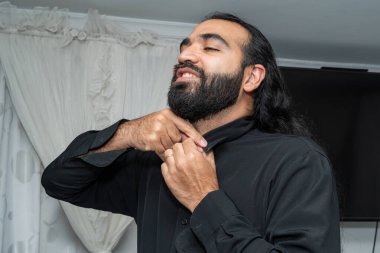 Arka planda televizyon olan beyaz perdelerin önünde düğme yakalı biri..