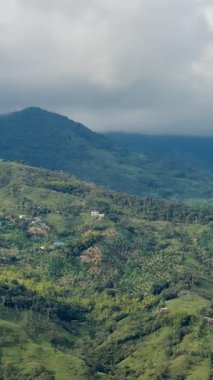 Kolombiya 'daki Cocorn dağlarının ve ekinlerin panoramik bulutlu manzarası, tepelerin arasında şehrin hava görüntüsüyle sona eriyor.