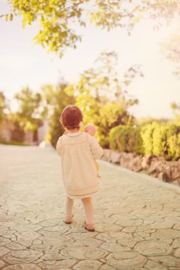 Arka toddler kız, başında çiçek çelenk ile parlak bir elbise giymiş ve bir bebek tutan. Küçük kız çiçekler ve ağaçlar ile bir bahçe de bir taş sokakta yürüyor. Güneş, güneş sisi, 