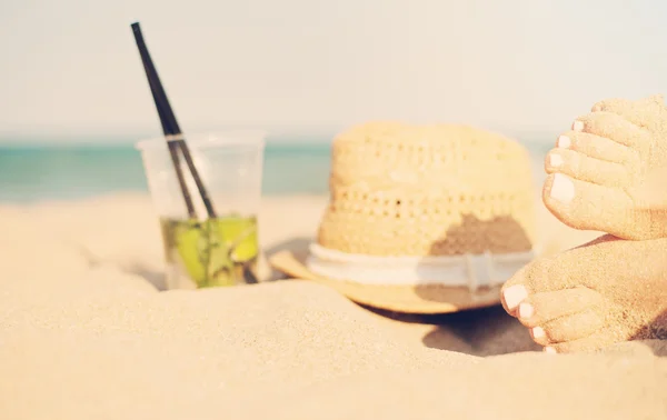 Ocio en verano - Hermoso de pies de mujer sexy, piernas femeninas en la playa de arena con sombrero y cóctel mojito. Cóctel mojito en la playa y sombrero de paja. Sol, sol neblina, resplandor — Foto de Stock