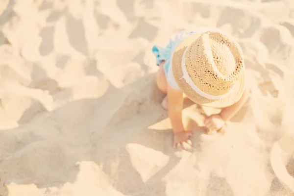 Niña niña con sombrero de paja y vestido azul jugando con arena en la playa en verano. Niña sentada en la orilla del mar. Sol, sol neblina, resplandor — Foto de Stock