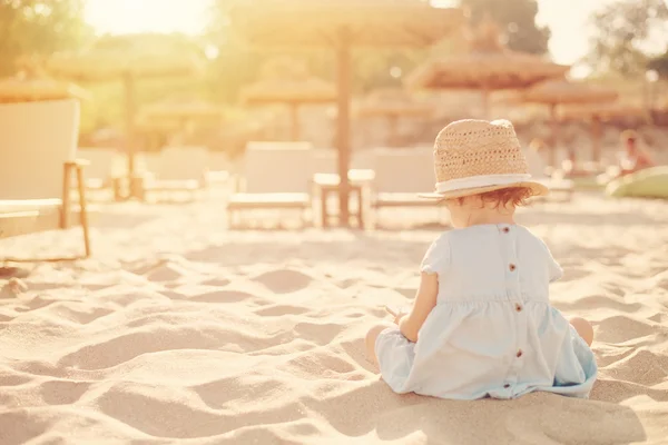 Bebé niña con sombrero de paja y vestido azul mirando el teléfono en un día soleado. Niña sentada en la orilla del mar. Sol, sol neblina, resplandor — Foto de Stock