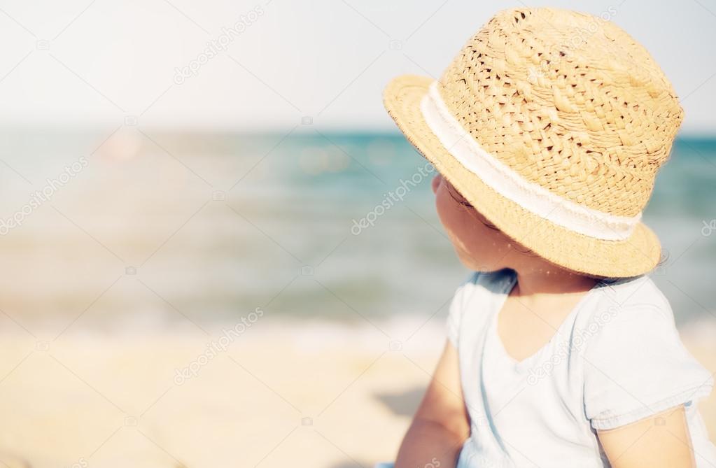 Baby girl child with straw hat and blue dress looking at the sea on a sunny day. Little girl sitting back on the shore of the sea. Sun, sun haze, glare