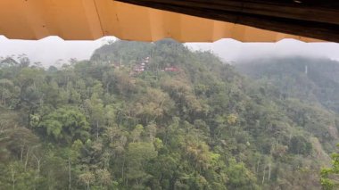 Puslu tepeler ve bereketli bir tropikal ormanın üzerine tünemiş küçük bir köy, kapalı bir balkondan hafif yağmurla görülüyordu..