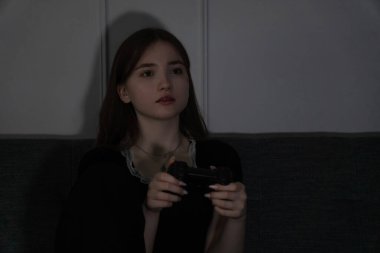 Genç bir kız bir kanepede oturur ve bir video oyunu oynamaya çok odaklanır. Loş ışıklandırma odası ve onun konsantrasyonu oyun oynamanın büyüleyici deneyimini vurguluyor.