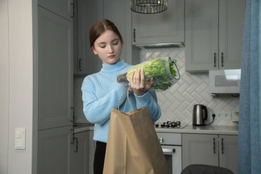 Yiyecek teslimatından sonra mutfaktaki kese kağıdından yiyecek çıkaran genç kız, masaya taze yeşil salata ve portakal suyu koyuyor.