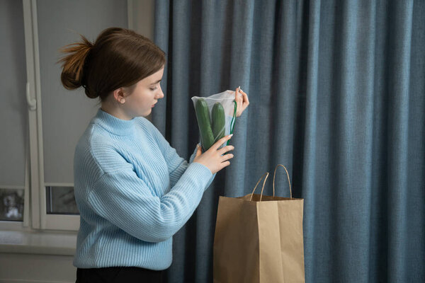 Young girl is unpacking reusable mesh bag with fresh cucumbers at home, standing next to paper bag and blue curtains, promoting sustainable lifestyle and zero waste concept