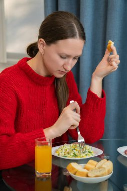 Kırmızı kazaklı, sağlıklı bir salata, kabuklu ekmek ve rahat evinde cam bir masada portakal suyu içen, dengeli bir yaşam tarzını kucaklayan genç bir kadın. Dikey fotoğraf