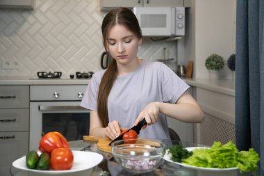 Genç kız ahşap bir tahtada taze domates kesiyor, şık, modern bir mutfakta akşam yemeği için renkli bir sebze salatası hazırlıyor, sağlıklı beslenme alışkanlıklarını kucaklıyor.