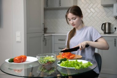 Genç bir kadın cam mutfak masasında oturmuş ahşap kesme tahtasındaki kırmızı dolmalıkları dikkatlice dilimliyor. Etrafı taze marul, dereotu ve diğer salata malzemeleriyle çevrili. Sağlıklı bir yemek hazırlıyor.