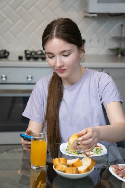 Young girl multitasking during breakfast, using a smartphone at the table. She is engaging with her device while having a meal, highlighting modern technology's role in daily life