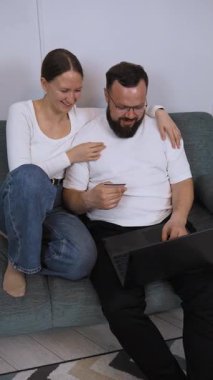 Neşeli çift rahat bir kanepeye uzanıyor, dizüstü bilgisayarlarını paylaşıyor ve kredi kartı kullanırken, aynı zamanda birlikte dikey video alışverişi yapıyorlar.
