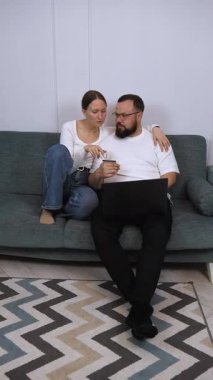 Genç çift evdeki rahat bir kanepede rahatlarken, online alışveriş için dizüstü bilgisayar kullanıyorlar ve ellerinde kredi kartı tutuyorlar, uygun ve modern bir perakende deneyimin keyfini çıkarıyorlar.