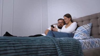 Genç çift yatakta yan yana yatar, akıllı telefonlara göz atar ve samimi dijital anı yatak odasında paylaşır, çağdaş ilişki bağlantısını temsil eder.