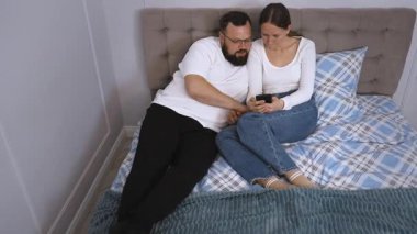 Genç çift yatakta yan yana yatar, akıllı telefonlara göz atar ve samimi dijital anı yatak odasında paylaşır, çağdaş ilişki bağlantısını temsil eder.
