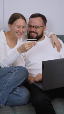 Genç çift evdeki rahat bir kanepede rahatlarken, online alışveriş için dizüstü bilgisayar kullanıyorlar ve ellerinde kredi kartı tutuyorlar, uygun ve modern bir perakende deneyimin keyfini çıkarıyorlar.