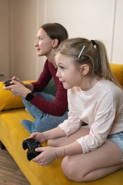 Konsantre olmuş kız ve anne video oyunu oynarken birbirlerine bağlanıyorlar, kumandayı tutuyorlar ve sarı bir kanepede oturup evde boş vakitlerinin tadını çıkarıyorlar. Dikey fotoğraf