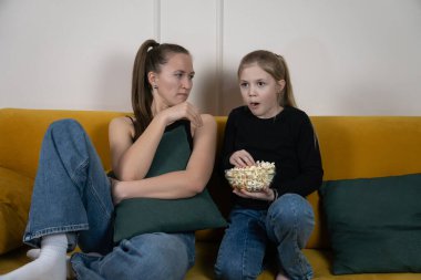Sarı bir kanepede oturan genç anne ve kız ellerinde patlamış mısırla korku ve şaşkınlıkla birlikte şüpheli bir film izliyorlar.