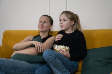 Genç bir kadın ve çocuk hardal sarısı bir kanepede birlikte oturuyorlar, bir ekran izliyorlar, bir kase patlamış mısırı paylaşıyorlar ve rahatlamış aile bağlarının tadını çıkarıyorlar.