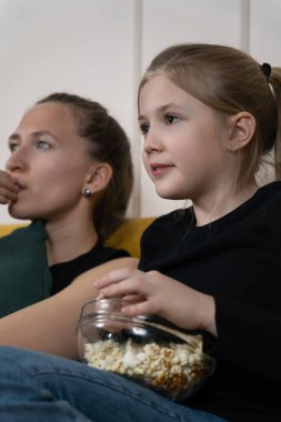 Anne ve kızı rahat bir film gecesinde kaynaşıyor, kanepede birlikte oturuyor, eğlence için televizyon izlerken bir kaseden patlamış mısır yiyorlar. Dikey fotoğraf
