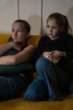 İki kız, sarı bir kanepede oturup ciddi ifadelerle televizyon seyrediyor. Biri yastığa sarılıyor, diğeri de loş bir odada elinde patlamış mısır tutuyor. Dikey fotoğraf