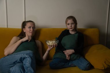 Anne ve kızı evde sarı bir kanepede dinleniyor, bir dizi izliyor ve televizyon programına ilgisizliklerini dışa bakarak ve patlamış mısır tutarak ifade ediyorlar.