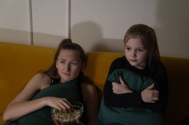 İki genç kız, loş ışıklı bir odada sarı bir kanepede oturmuş, yoğun bir şekilde korku filmi izliyorlar, biri patlamış mısır yiyor, diğeri de yastık tutuyor, korku ve gerilim ifadeleri sergiliyorlar.