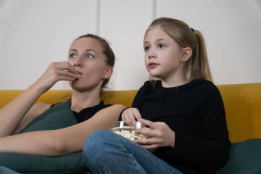 Genç bir kadın ve kız kanepede oturuyor, birlikte televizyon izliyorlar, bir kase patlamış mısır paylaşıyorlar ve evde rahat bir aile tatili geçiriyorlar.