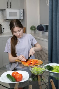 Genç bir kız ahşap kesim tahtasında dolma biber doğruyor, sağlıklı vejetaryen salatası yapıyor ya da modern mutfakta yemek pişirmek için malzemeler hazırlıyor. Dikey fotoğraf