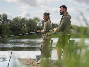 Nehir kıyısında duran genç çift, ellerinde olta kamışlarıyla, doğanın güzelliği ve suyun sükuneti ile çevrili, boş bir günün tadını çıkarıyorlar.