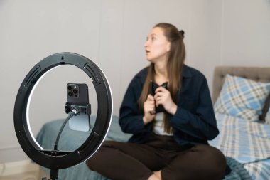 Nüfuzlu genç bir kadın akıllı telefonuyla bir video kaydetmeye hazırlanırken halka ışığını ayarlıyor, evinde yatağında rahatça oturuyor, içerik yaratma işine dalmış..