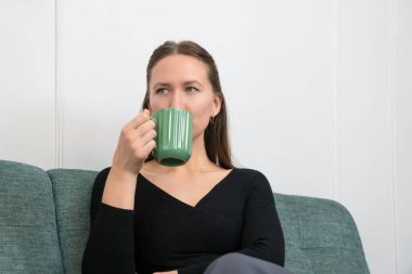 Genç bir kadın rahat bir kanepede oturmuş sıcak bir içeceğin tadını çıkarıyor, derin derin bakıyor, rahat ve düşünceli bir atmosfer yaratıyor.