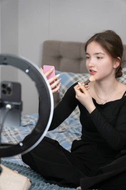 Ergen, yatağında otururken makyaj yapıyor ve video kaydediyor. Yüzük lambası ve akıllı telefon kullanarak. Küçük bir aynada çalışıyor. Modern güzelliğin ve kendini ifade etmenin bir örneği.