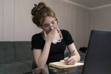 Yorgun bir kız öğrenci ya da serbest çalışan, elini uzatıp ağzını kapatırken deftere notlar yazıyor, çalışıyor ya da uzaktan çalışıyor.