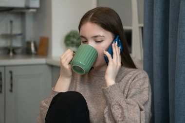 Young girl enjoys a relaxing moment at home, sitting comfortably while sipping tea from a green mug and engaging in a phone conversation
