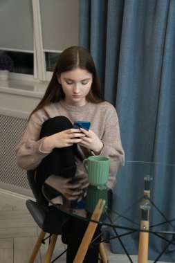 Yoğunlaşmış genç kız evde akıllı telefon kullanıyor bir fincan kahve ya da çayla cam masada oturuyor, günlük kıyafetler giyiyor, internette veya sosyal medyada geziniyor. Dikey fotoğraf