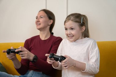 Anne ve kızı renkli bir kanepede mutlu bir şekilde oturup eğleniyorlar. Kumandanlarla video oyunları oynayıp gülüyor ve eğleniyorlar.