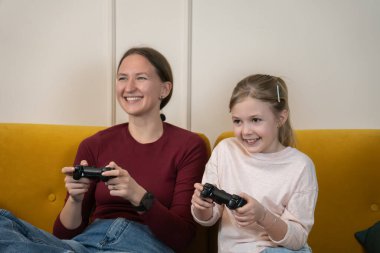 Gülümseyen anne ve küçük kızı video oyununun keyfini çıkarırken, evdeki rahat sarı koltukta birlikte otururken, mutlu ve mutlu bir aile eğlencesi anı paylaşıyorlar.