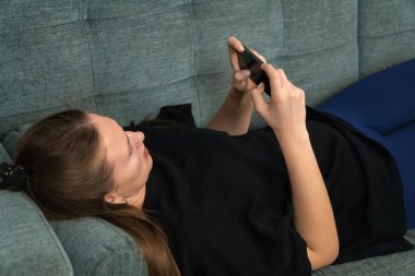 Uzun kahverengi saçlı genç bir kadın, siyah bir tişört giyiyor, rahat bir kanepede rahatlarken akıllı telefonunu karıştırıyor, oturma odasında boş zamanlarının keyfini çıkarıyor.